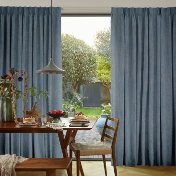 Blue pleated full-length curtains, hung and drawn slightly apart to reveal a glass patio door, framing a sunlit backyard; dining table with pendant lamp, pancakes and wooden chairs in foreground.