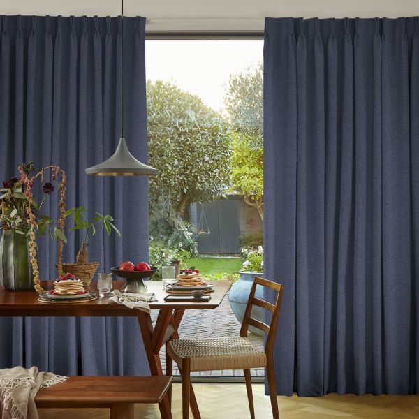 Floor-length navy-blue pleated curtains, drawn open at center to reveal a window; framing a sunlit garden beyond while a wooden dining table with pancakes and a pendant lamp fills the foreground.