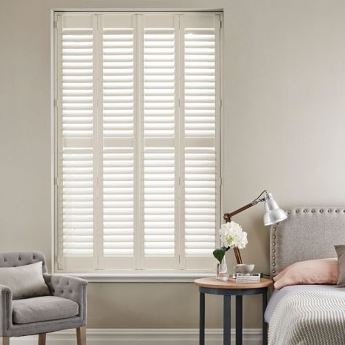 White plantation shutters, four hinged louvered panels covering the window, closed with slats angled to admit diffused light into a neutral bedroom corner with armchair, bedside table, lamp and flowers.