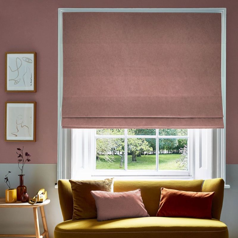 A textured dusty-pink Roman blind is lowered over a white-framed sash window, partially filtering daylight in a living room with a mustard-yellow sofa, cushions, side table and green garden view.
