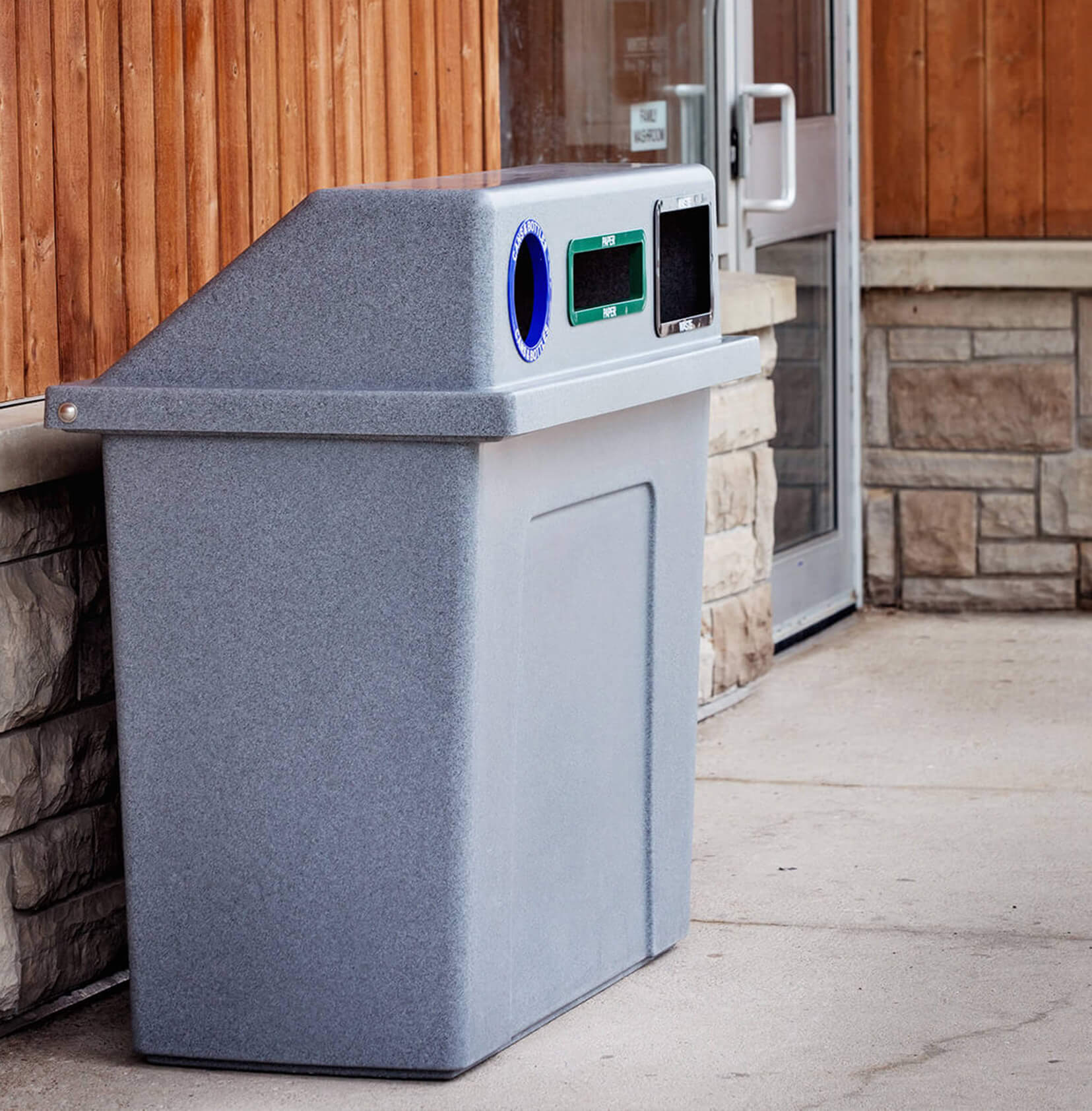 durable plastic trash and recyclables collection container with sloped hinged lid and color coded restrictive openings outside a municipal building