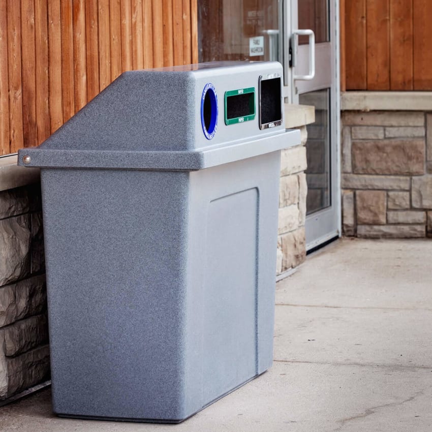 durable plastic trash and recyclables collection container with sloped hinged lid and color coded restrictive openings outside a municipal building