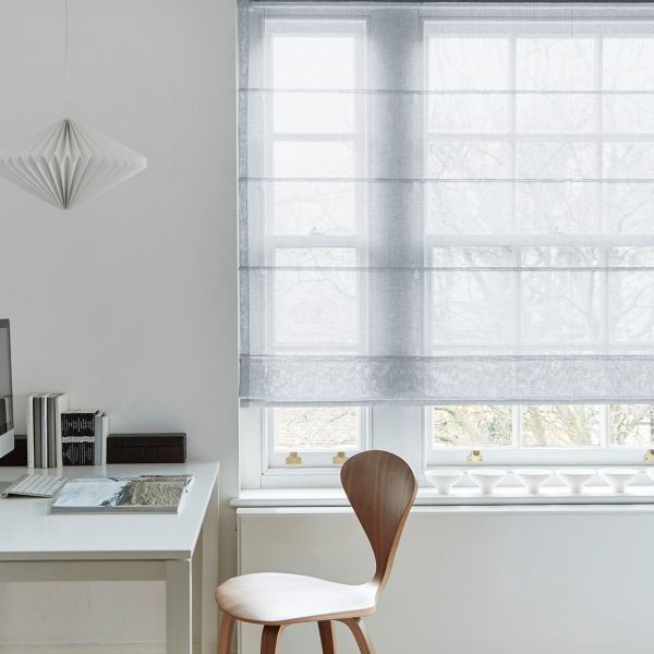 Semi-sheer, light-gray Roman shade with horizontal folds, lowered over a multi-pane sash window, diffusing pale daylight and obscuring leafless trees outside; minimalist home office with white desk, wooden chair.