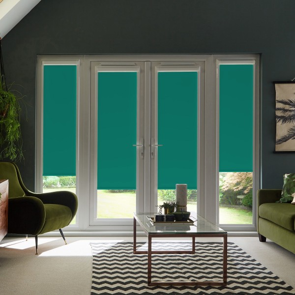 French doors with teal roller blinds, lowered to cover most panes, stand closed in a modern living room with green armchairs, glass coffee table on chevron rug and garden beyond.