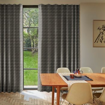 Dark curtains patterned with repeating geometric circles, hanging beside a tall window, partially drawn to reveal panes and filter daylight; dining area with wooden table, chairs and garden view.