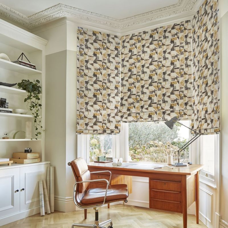 Patterned Roman blinds covering a bay window â€” geometric beige, gray and mustard print â€” partially lowered, filtering soft daylight; home office with mid-century wooden desk, leather swivel chair and built-in shelves, garden view.