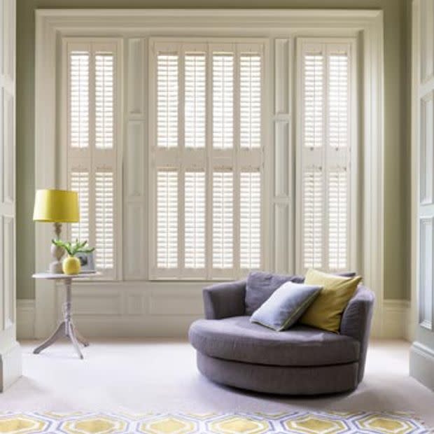White plantation shutters cover a bay window, closed and filtering soft daylight; a round gray chair with two pillows sits beside a small side table topped by a yellow lamp.