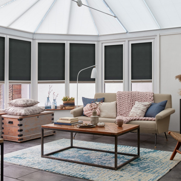 Dark grey cellular pleated blinds covering bay conservatory windows, lowered to mid‑height and blocking light; sunroom with beige sofa, chunky pink throw, wooden coffee table on blue rug.
