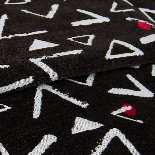 Black knit throw patterned with white, rough-edged triangles and occasional red semicircles; folded in overlapping layers, displaying textured weave and seams in a close-up, softly lit studio view.