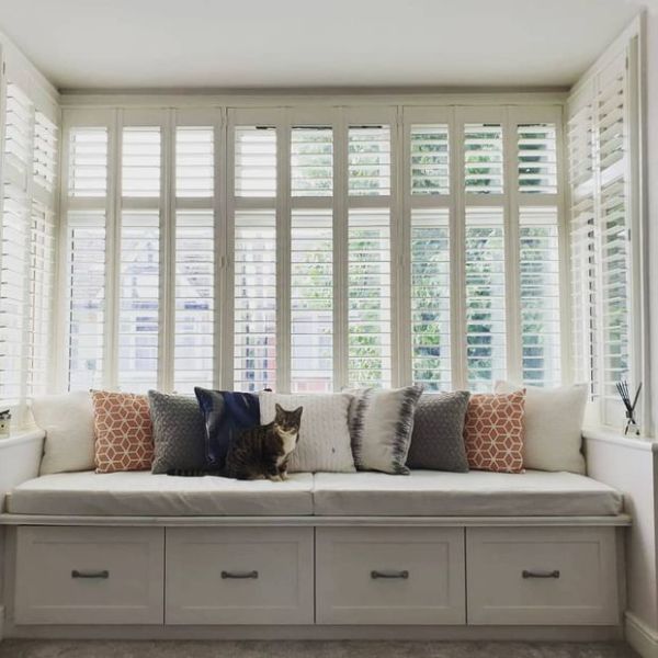 White plantation shutters cover tall bay windows, slatted and partially open to filter daylight; a built-in cushioned bench with patterned pillows and a tabby cat sits in a bright, airy room.