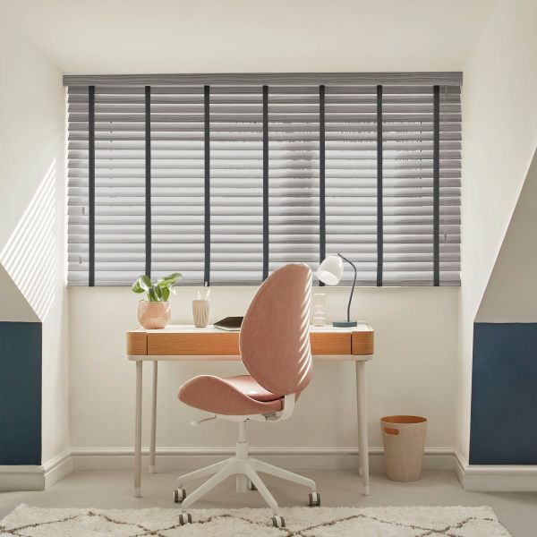 Gray horizontal slatted blinds with dark vertical tapes over a window, mostly closed, filtering daylight and casting stripes across an attic study with wooden desk, pink swivel chair and lamp.