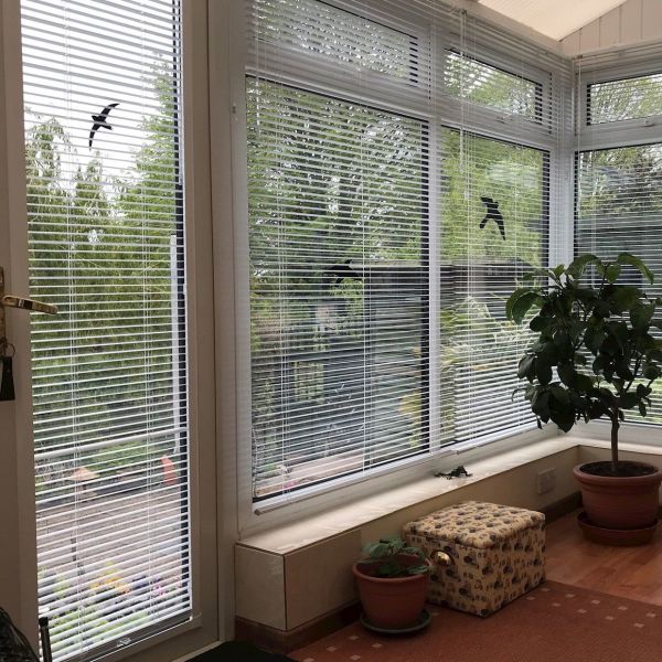White horizontal Venetian blinds cover tall bay windows and a glass door, with black bird-silhouette stickers, partially closed and filtering daylight; conservatory with potted plants, patterned ottoman and garden view.