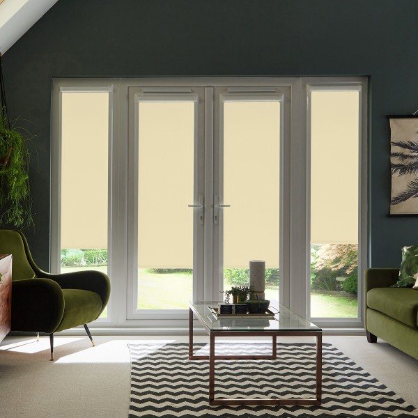 French glass doors with beige roller blinds are closed, filtering daylight into a living room with green armchairs, a glass coffee table on a chevron rug and potted plants.