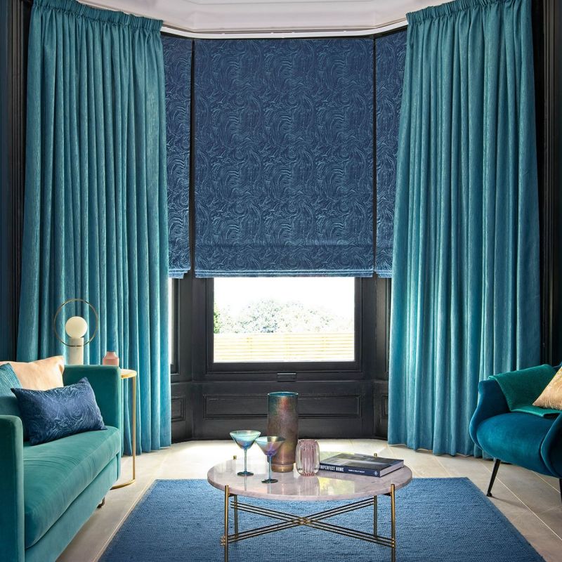 Teal floor-to-ceiling velvet curtains frame a lowered patterned navy Roman blind covering most of a bay window, filtering daylight. Cozy living room with teal sofas, marble coffee table, garden view.

IMPERFECT HOME