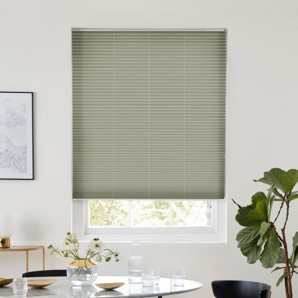 Sage-green pleated blind, fully lowered over a window with visible lower sash, filtering daylight; above a dining table with glassware, vase of daisies, framed art and a fiddle‑leaf fig.