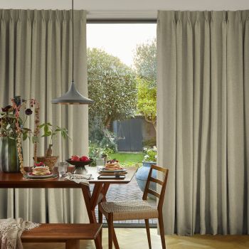 Full-length beige pleated curtains, drawn partly aside to reveal a central opening, framing and filtering soft daylight over a dining table set for breakfast and a green garden with trees and shed.