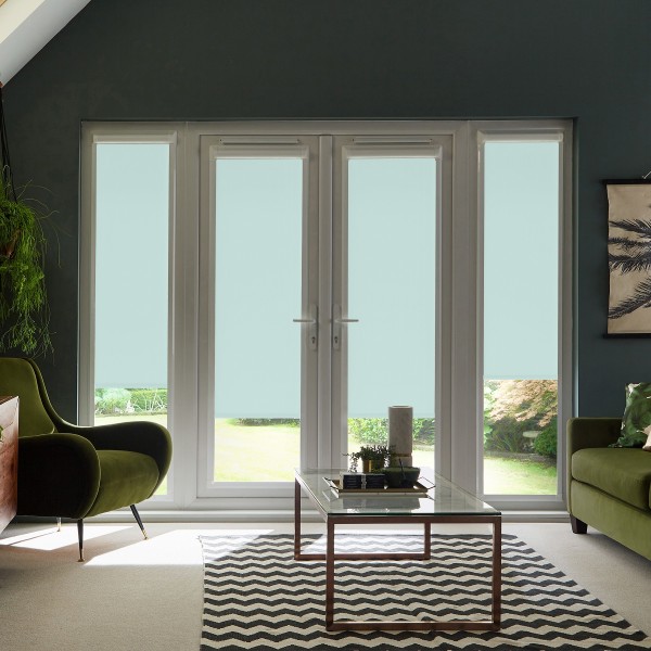 Glass patio doors with pale blue roller blinds covering the lower panes, framing a garden outside; flanked by green armchairs and a glass coffee table on a chevron rug in a modern living room.