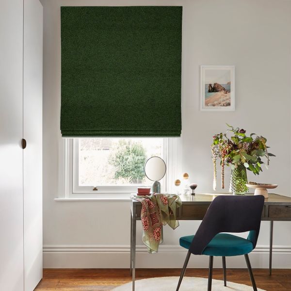 Dark green Roman blind, fully lowered with neat horizontal folds, blocking light; bright home office with wooden desk, teal chair, vase of flowers and outside garden visible.