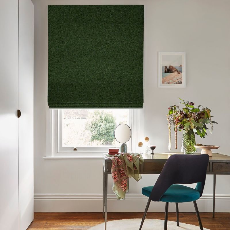 Dark green Roman blind, fully lowered with neat horizontal folds, blocking light; bright home office with wooden desk, teal chair, vase of flowers and outside garden visible.