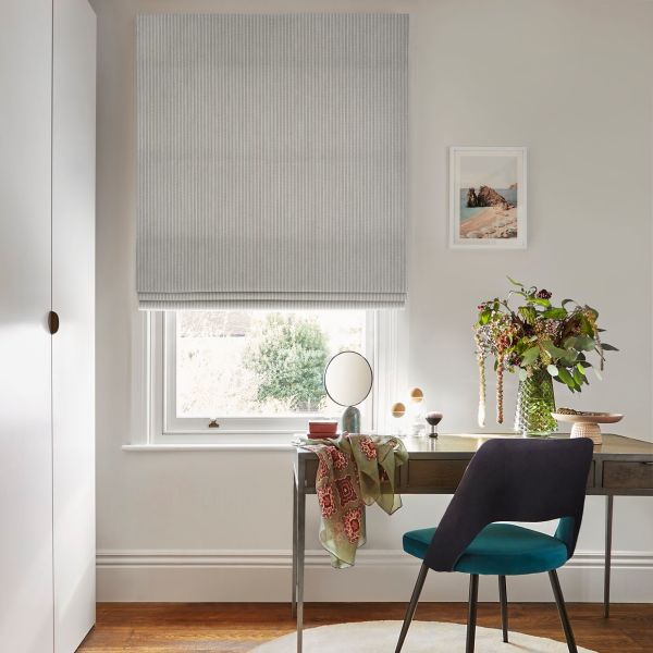 Grey textured Roman blind lowered across the window with soft folds, filtering daylight; a desk holding mirror, vase of flowers and teal chair in a neutral study overlooking a garden.