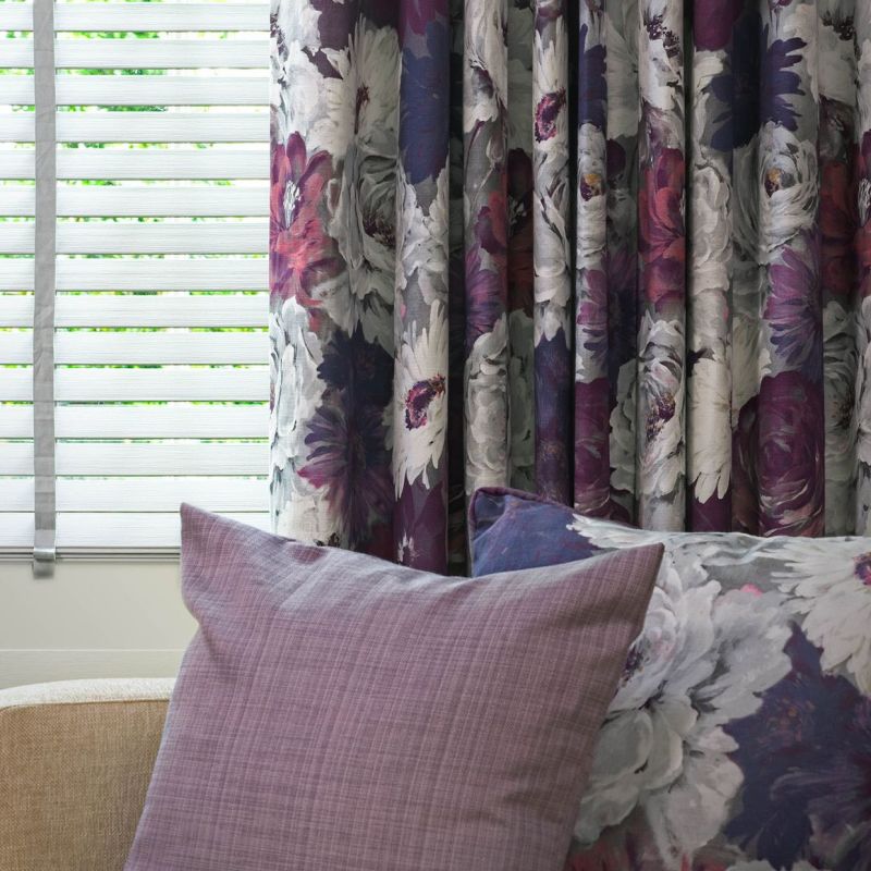 Floral curtains (purple, white peony pattern) hang pleated on the right, partially covering white horizontal blinds filtering daylight; mauve and floral cushions on nearby seating in a softly lit room.