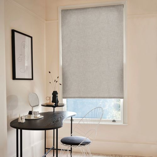 Light-gray fabric roller blind, fully lowered over a tall window, subtly diffusing daylight; simple home desk nook with black curved table, wire chair, framed line-art, small vase and natural light.