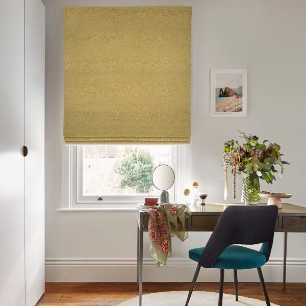 Textured mustard-yellow Roman blind, lowered and filtering daylight over a white sash window above a wooden desk with teal chair, vase of flowers, framed seaside print, soft natural light.