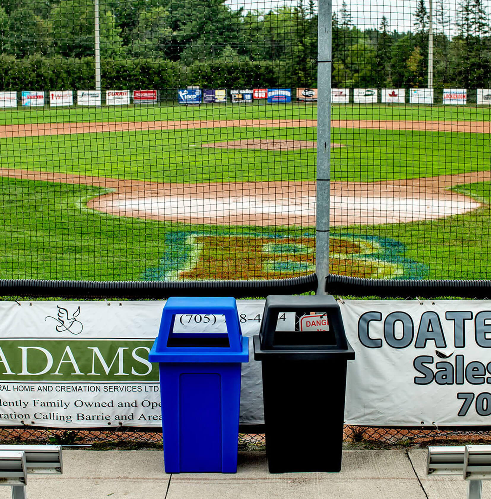 outdoor blue plastic recycling bin beside a black waste bin behind home plate at a baseball field