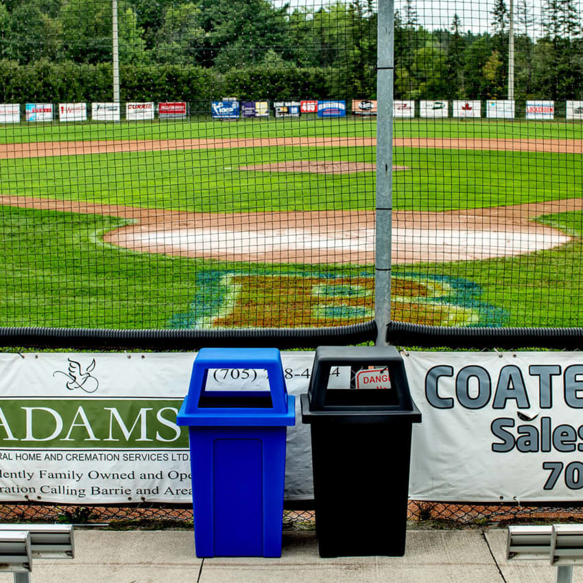 outdoor blue plastic recycling bin beside a black waste bin behind home plate at a baseball field