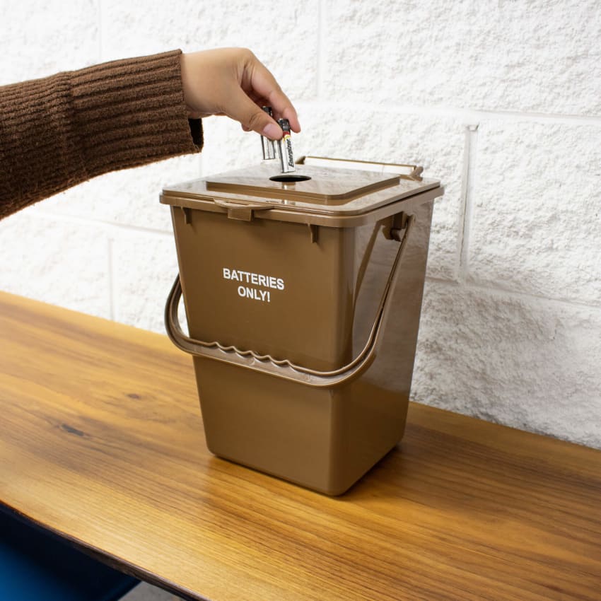 brown plastic container for collecting used battering for recycling placed on a wood shelf with a person depositing batteries