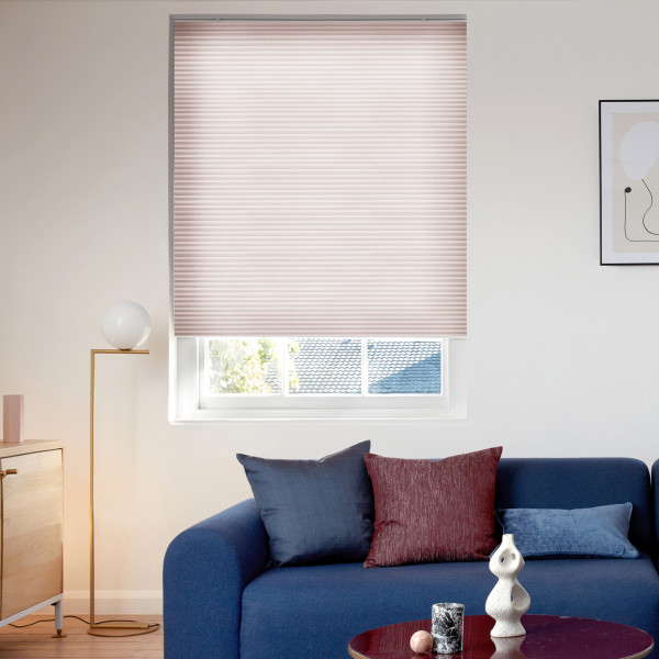 Light pink pleated cellular blind, fully lowered covering the window, gently filtering daylight; modern living room with blue sofa, colored cushions, floor lamp, coffee table and framed art, rooftop view below.