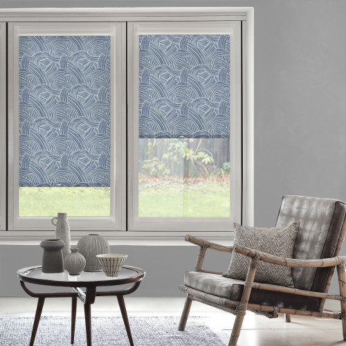 Blue roller blinds with white wave pattern cover twin windows' upper halves, lowered partially and filtering light; daylight living room with gray walls, rattan armchair, cushion, table and lawn view.