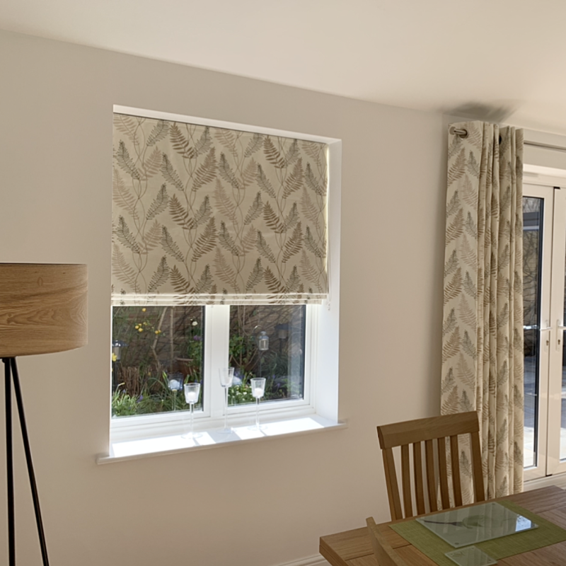Patterned roller blind and matching grommet-top curtain, beige with fern print, lowered over a sash window (blind half-down) filtering daylight; dining area with wooden table, chair, lamp and garden view.