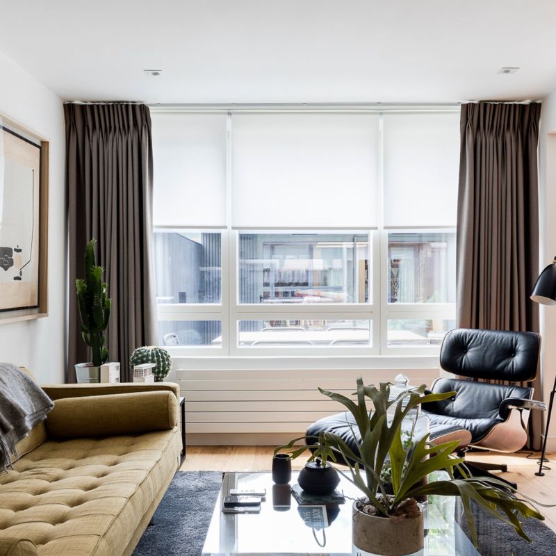 Brown pleated side curtains and lowered white roller blinds framing a three-panel window, blinds diffusing daylight; living room with mustard sofa, black leather lounge chair, plants and glass coffee table.