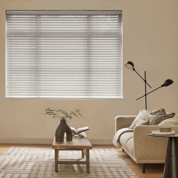 White horizontal blinds covering a window, closed and filtering daylight; beige living room with a low wooden coffee table, sculptural vase and open book, sofa and adjustable floor lamp.