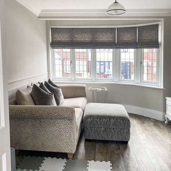 Gray fabric Roman blinds across a bay window, partially lowered and filtering daylight; living room with beige patterned corner sofa, gray ottoman, round side table, wood floor and street view.