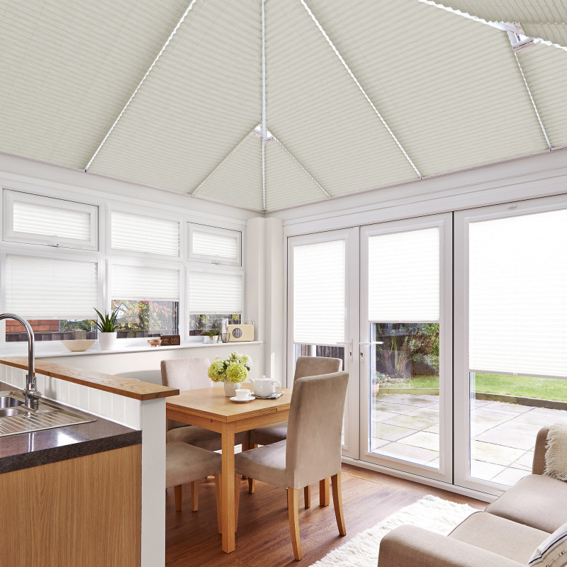 Cream pleated cellular blinds fitted across the pitched conservatory roof and over windows and French doors, lowered to diffuse bright daylight; dining-kitchen conservatory with wooden table, chairs, and garden view.