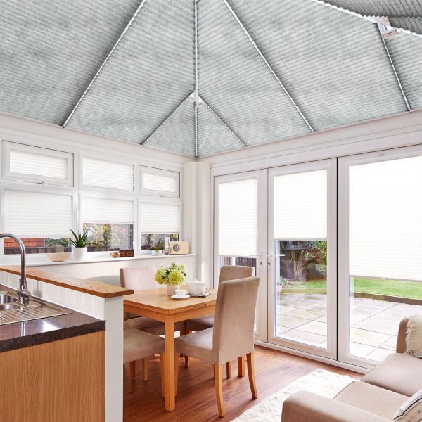 Pleated cellular blinds on the pitched conservatory roof and matching white pleated blinds on windows and patio doors, mostly lowered and filtering soft daylight; dining area opening onto a garden.