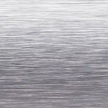 Brushed stainless-steel surface with fine horizontal grain; reflecting diffuse light and subtle highlights, close-up view suggesting a sheet-metal or appliance panel in a neutral, evenly lit studio context.
