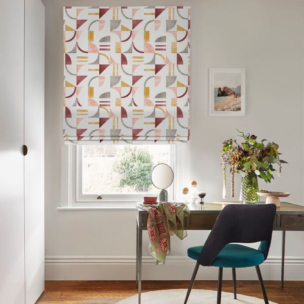 Geometric-patterned Roman blind (pink, mustard, grey, burgundy) lowered and gently folded, filtering daylight over a home desk with teal chair, mirror, vase of flowers and framed seaside print.