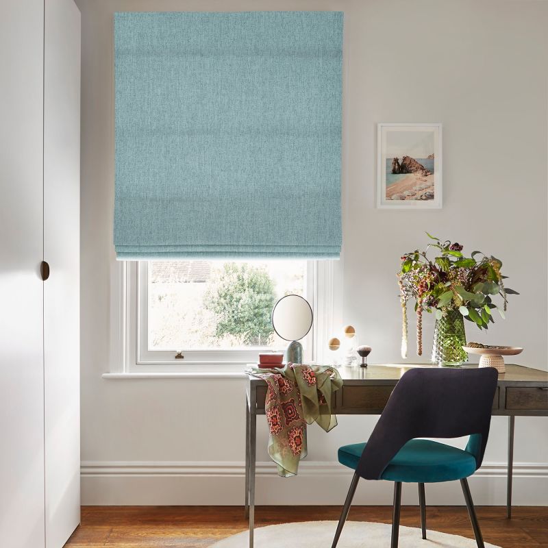 Light teal fabric Roman blind, fully lowered with horizontal folds, gently filtering daylight; home office with wooden desk, teal chair, vase of flowers and window view of trees.