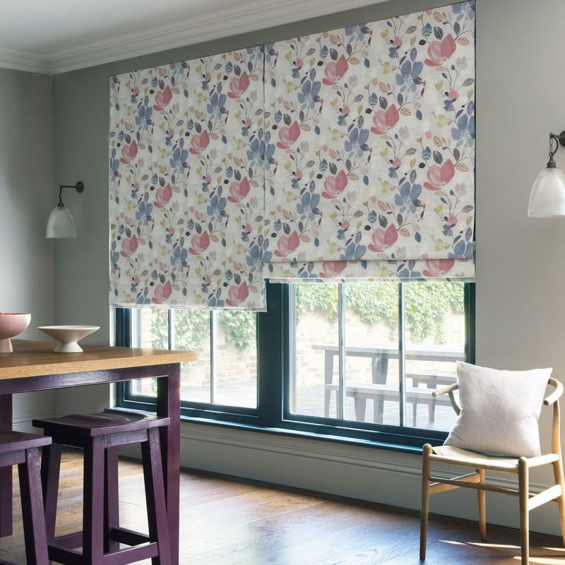 Floral Roman blind, lowered with pleats and pink-blue botanical print, filtering daylight over a black-framed window in a bright dining nook with wooden table, purple stools and a chair.