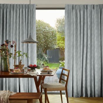 Floor-to-ceiling pleated grey curtains, hanging on either side of a sliding glass door, parted to reveal a sunlit backyard; interior dining table set with pancakes, pendant light and woven chair.