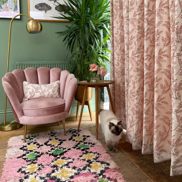 Full-length pink floral curtains at right, heavy fabric with botanical print; hung closed, softly filtering daylight; living-room corner with pink velvet chair, lamp, tall plant, shag rug, and cat.