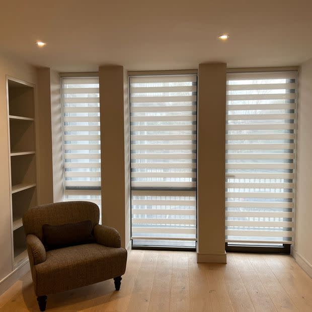 Zebra roller blinds covering three tall, narrow windows, with alternating opaque and sheer horizontal bands filtering soft daylight. Room: neutral living area with armchair, built-in shelves, recessed ceiling lights.