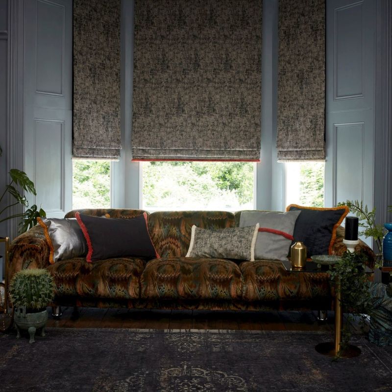 Three textured Roman blinds, lowered mid-height and filtering daylight; in front, a patterned tufted sofa with assorted cushions; indoor plants and side table in a softly lit living room.