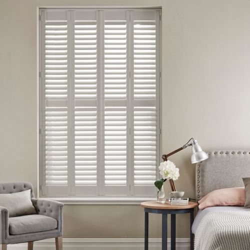 White full-height louvered plantation shutters covering a tall window, slats slightly tilted filtering soft daylight; bedroom setting with upholstered bed, bedside table and lamp, neutral walls and armchair.
