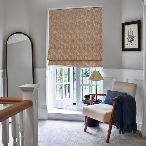 Patterned beige Roman blind, lowered and slightly folded at the bottom, filtering daylight over a ground-floor casement window opening to a garden, in a lit landing with armchair, lamp, mirror.
