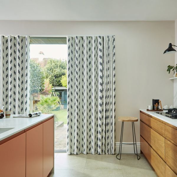Floor-length patterned white curtains with vertical charcoal brushstrokes, partially drawn across a sliding glass door, letting daylight and a backyard garden view into a bright modern kitchen with wooden cabinets.