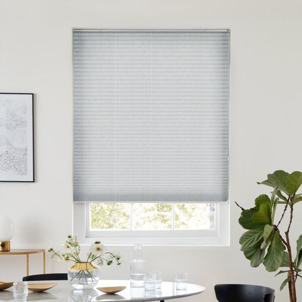Light-gray pleated cellular blind, fully lowered across the window with horizontal texture, filtering soft daylight into a dining nook with table, glassware, vase of flowers and a fiddle-leaf fig.